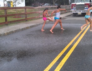 Young girls run barefoot in the street under a sprinkler.